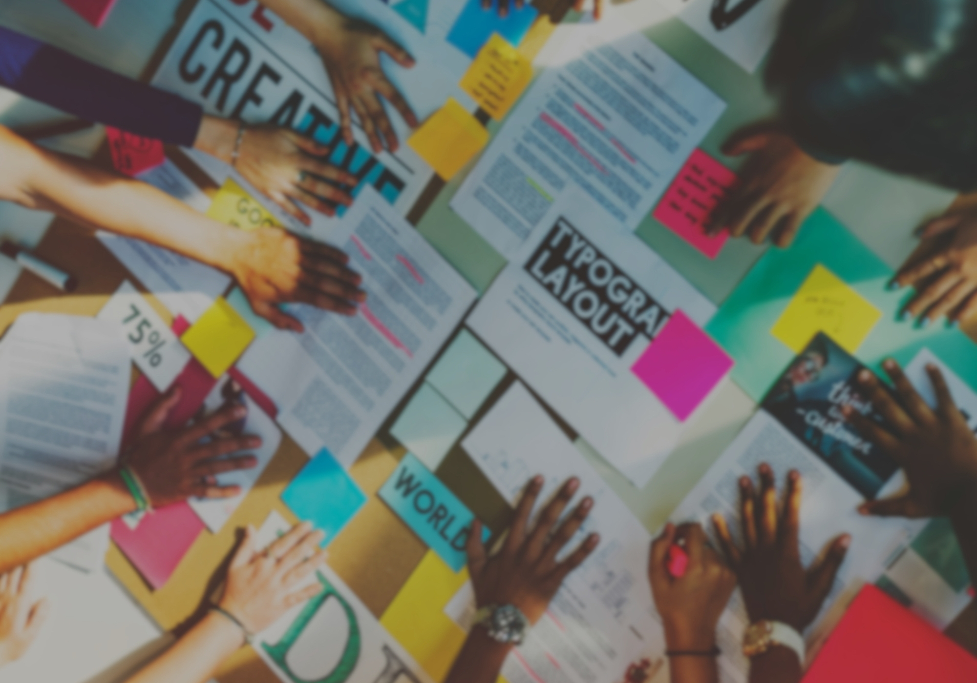 Overhead view of many hands arranging sticky notes, posters, and printed pages on a creative planning table.