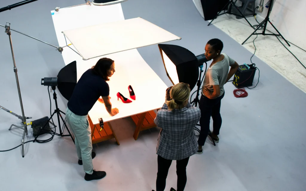 Behind-the-scenes view of a creative studio production team using overhead lights, softboxes, and a seamless white table setup to shoot red high heel shoes.