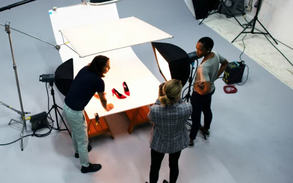 Behind-the-scenes view of a creative studio production team using overhead lights, softboxes, and a seamless white table setup to shoot red high heel shoes.
